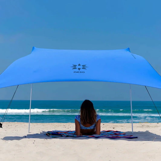A woman sits on a striped beach towel under a large blue Neso canopy on a sandy beach with the ocean and blue sky in the background.