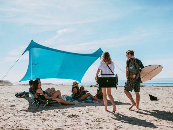 A bright blue beach canopy shade tent set up on a sandy beach with several people relaxing underneath and others walking nearby, including one person carrying a surfboard.