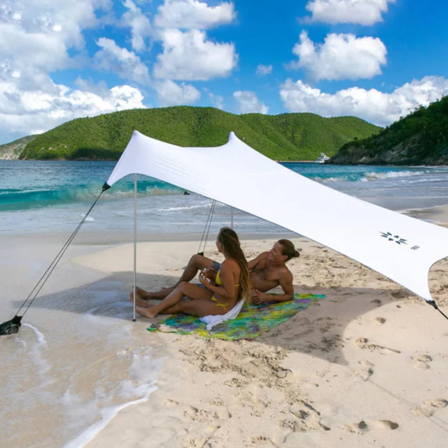 A white beach canopy tent is set up on a sandy beach with two people sitting underneath on a colorful towel, with ocean water, green hills, and a blue sky in the background.