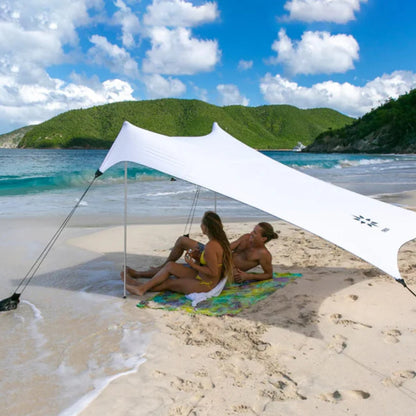 A white beach canopy tent is set up on a sandy beach with two people sitting underneath on a colorful towel, with ocean water, green hills, and a blue sky in the background.