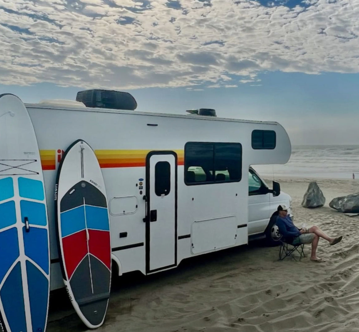 RV on a beach with surfboards and a person sitting nearby, featuring the Honu brand.