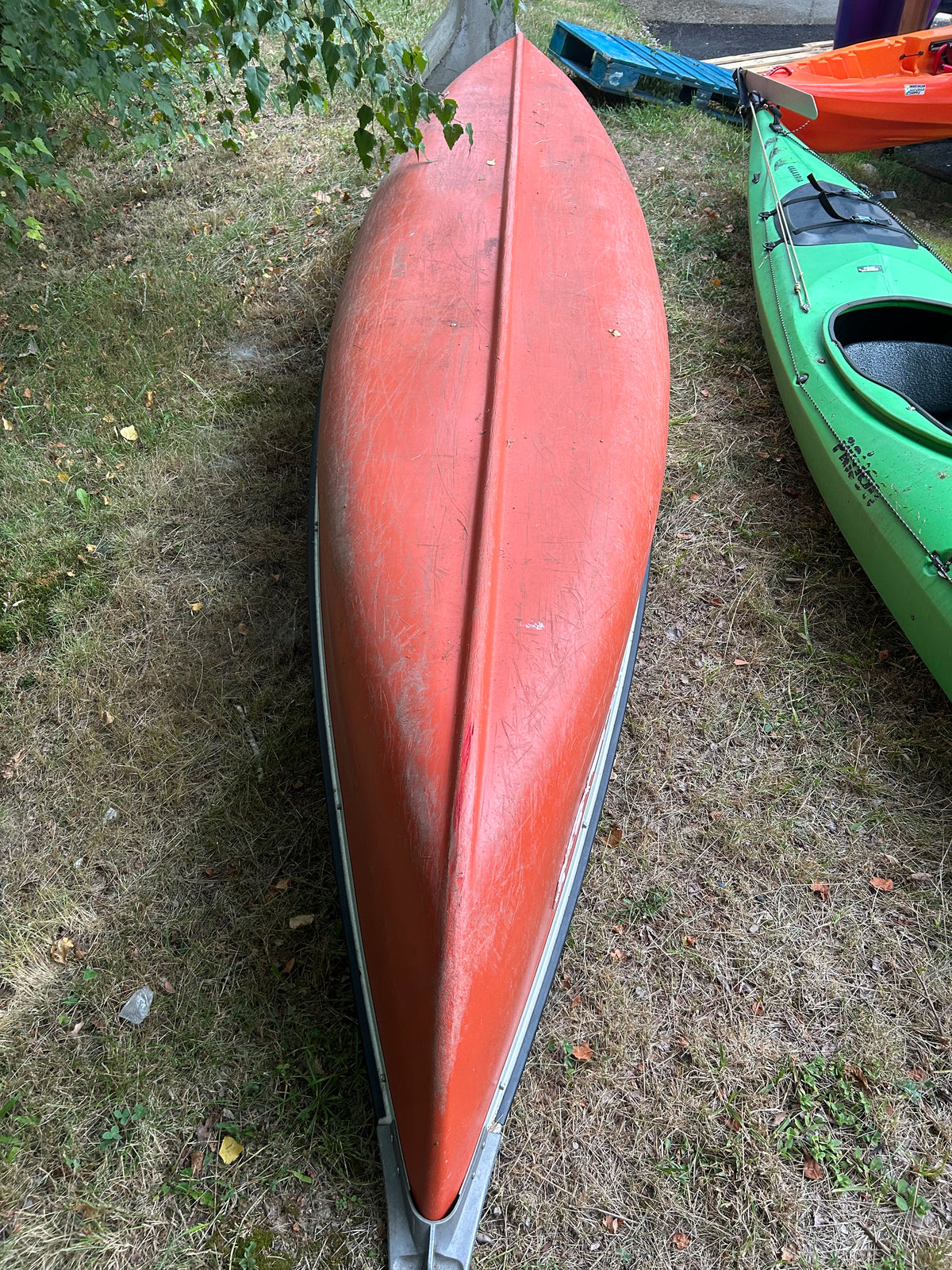 An orange-red canoe or kayak photographed outdoors on grass, with other boats partially visible at the right edge.